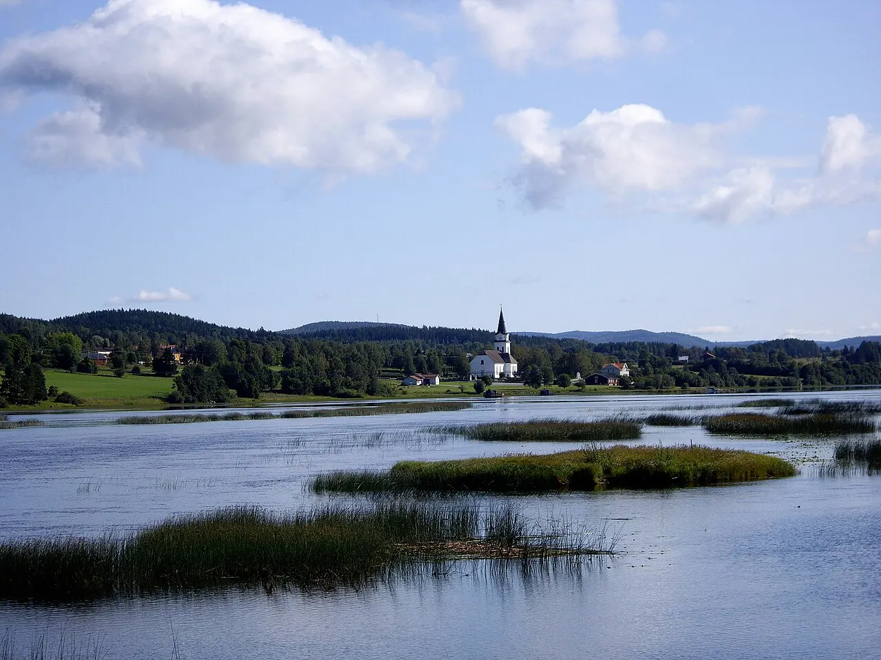 Fotografi av Torps kyrka i Fränsta, Sverige. Fotot är taget från andra sidan vattnet.